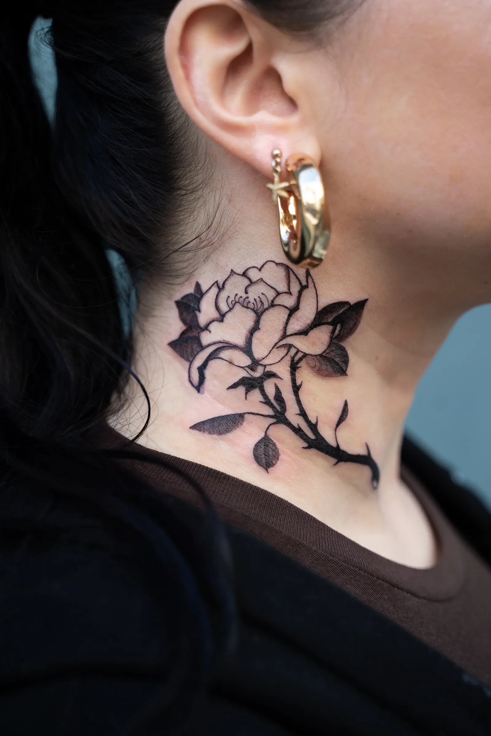 A peony on a thorny stem with leaves, tattooed in black and grey on the side of a neck
