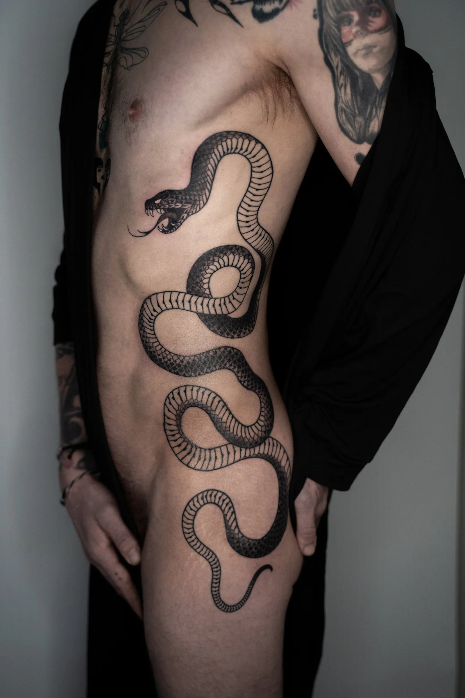 A large black snake winding down the side of a torso from the ribs to the hip, its mouth open near the chest