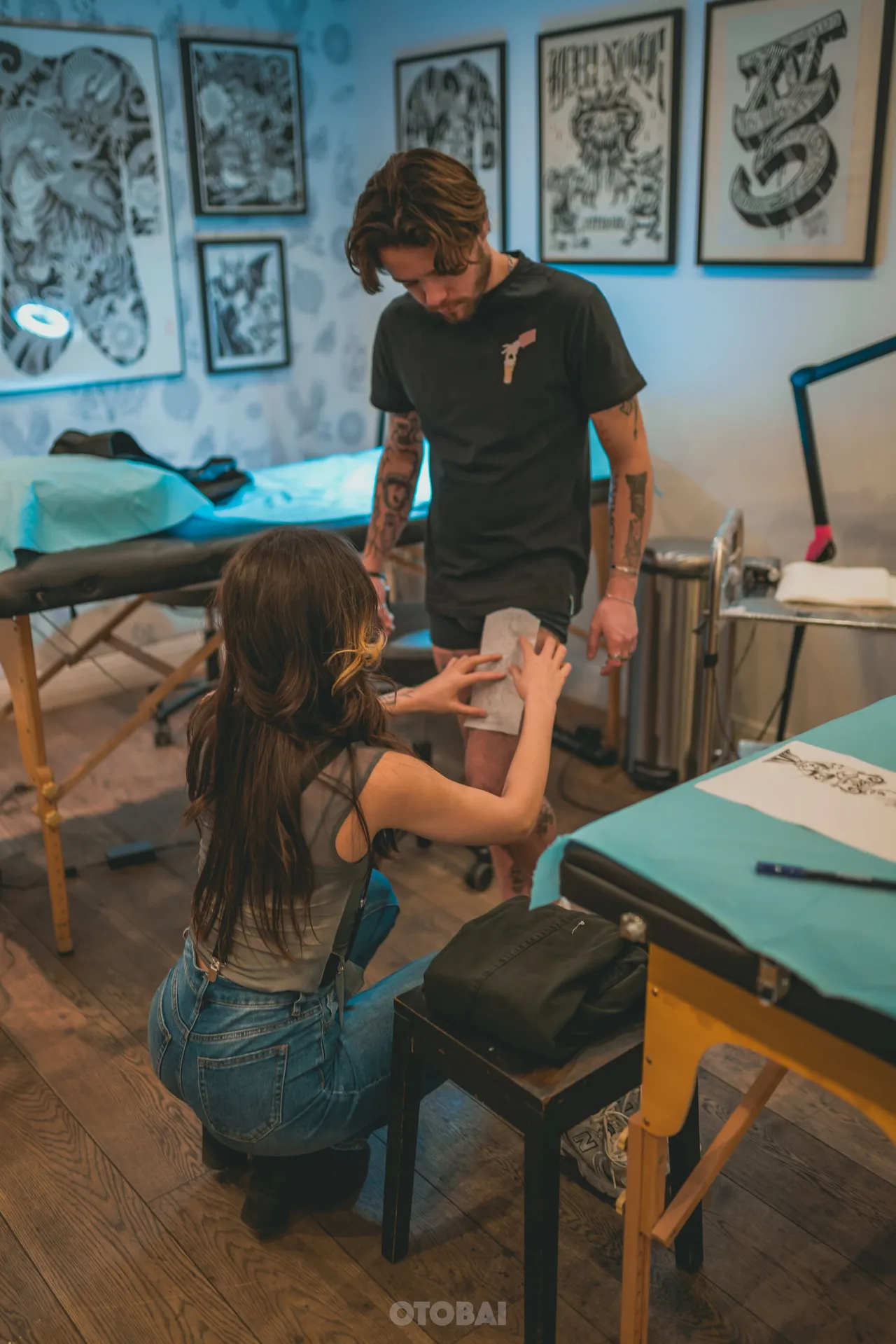 Joy kneeling to place a stencil on a client's leg, framed flash on the walls