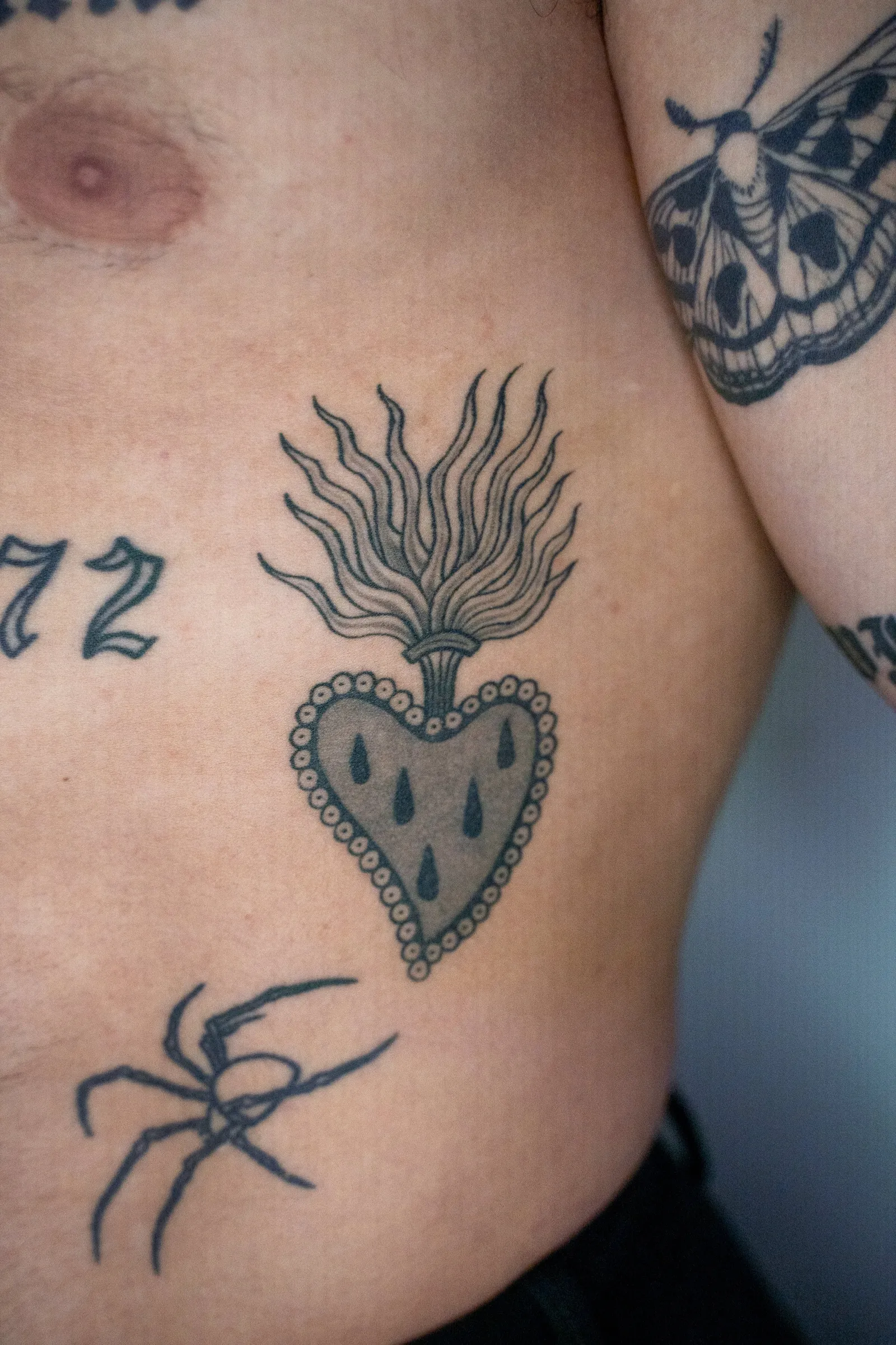 A flaming sacred heart with teardrops on the side of a torso, healed, with a spider and a moth nearby