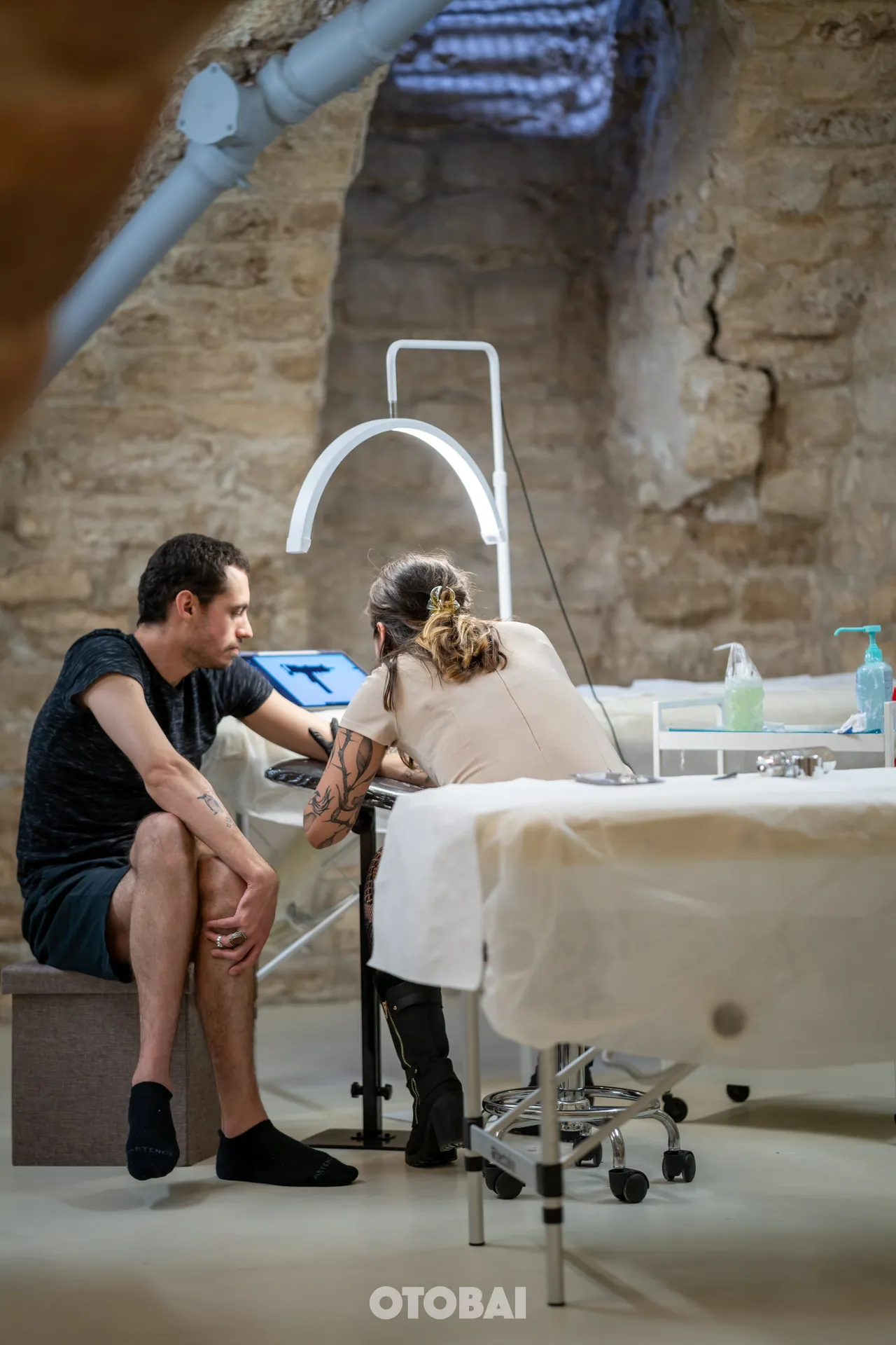 Joy tattooing a client's arm in a stone-walled room