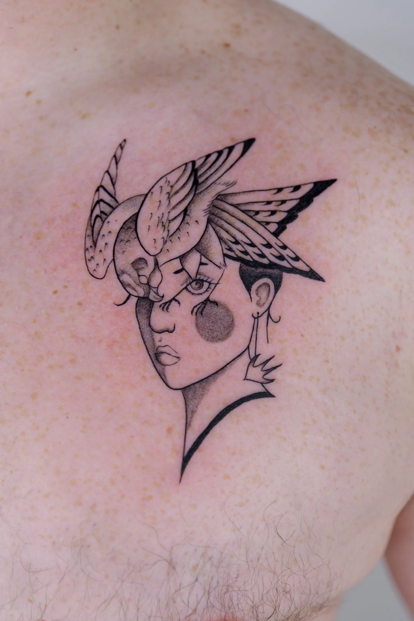 A woman's face crowned by a bird with spread wings, tattooed in black and grey on a chest