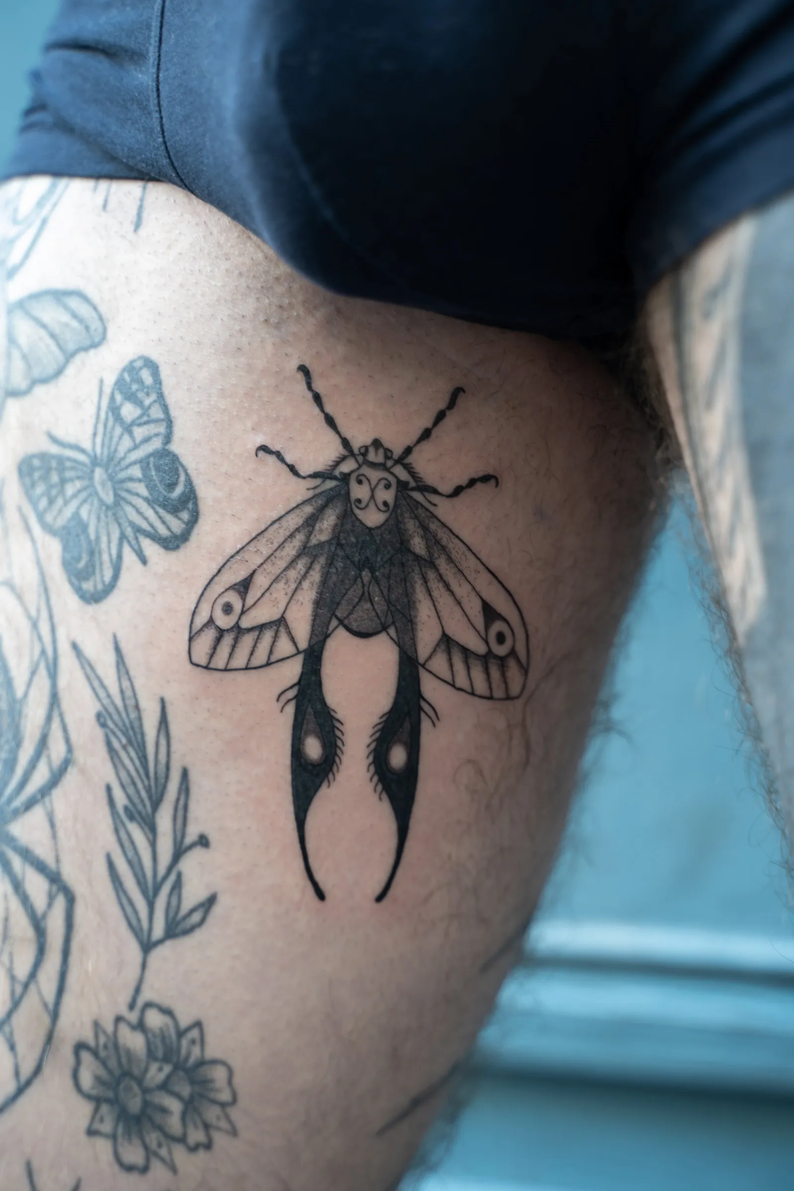 A moth with patterned wings and long trailing tails, tattooed on a thigh beside older tattoos