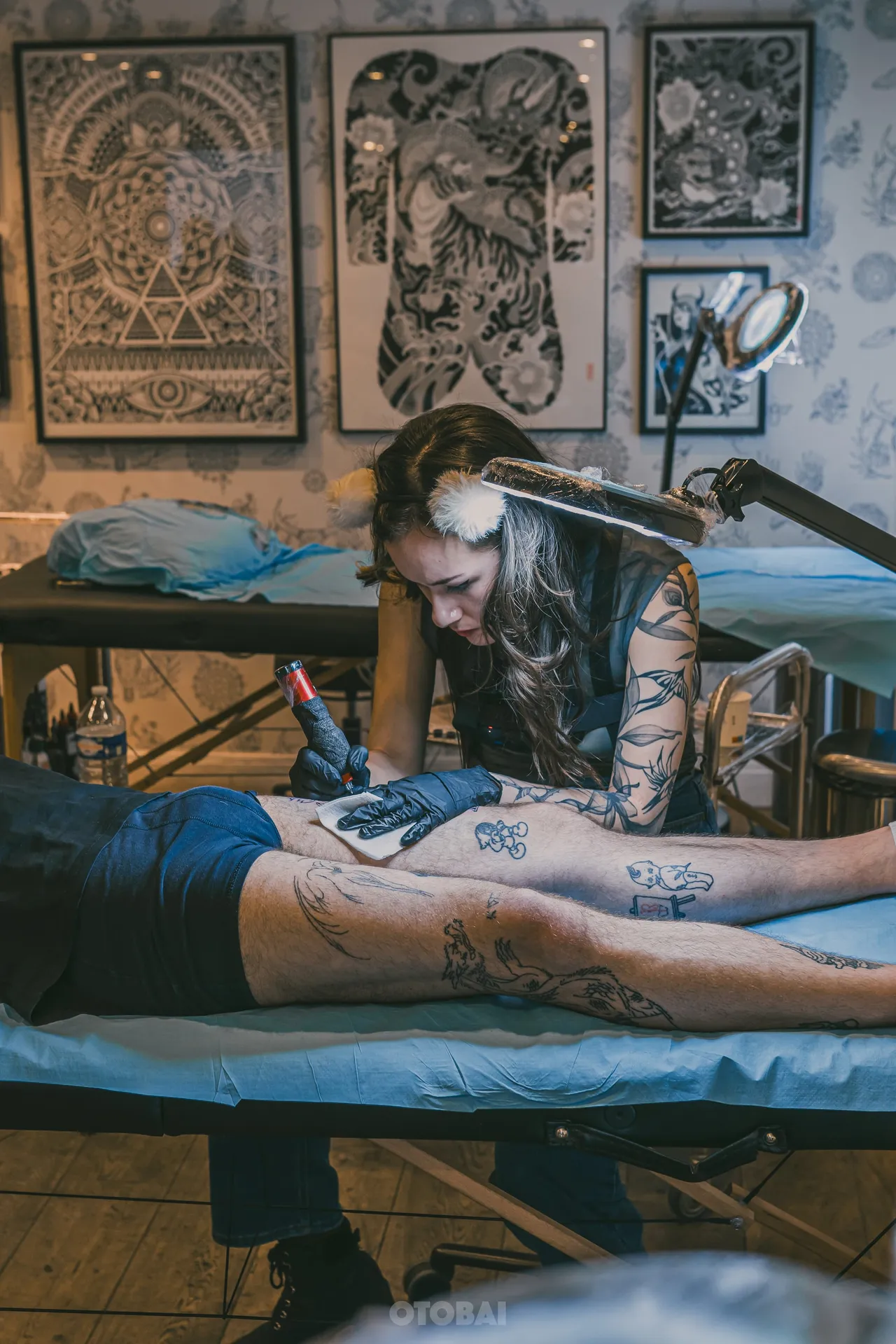 Joy tattooing a client lying on the table, framed flash on the wall behind her