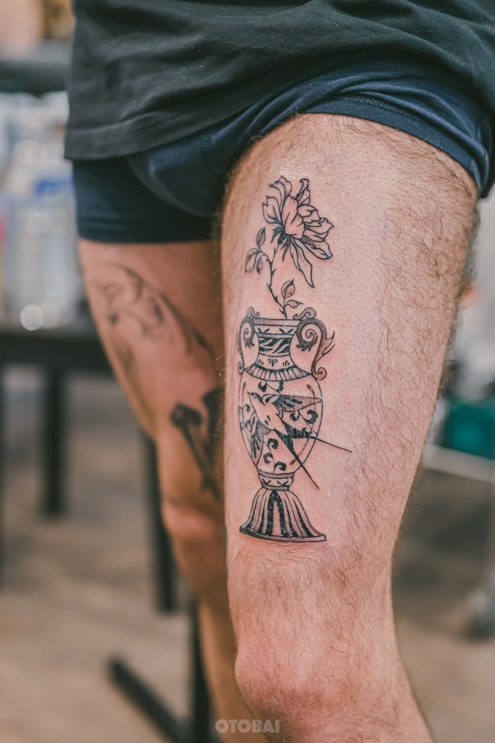 A decorated vase holding a full bloom, tattooed in black and grey on a thigh