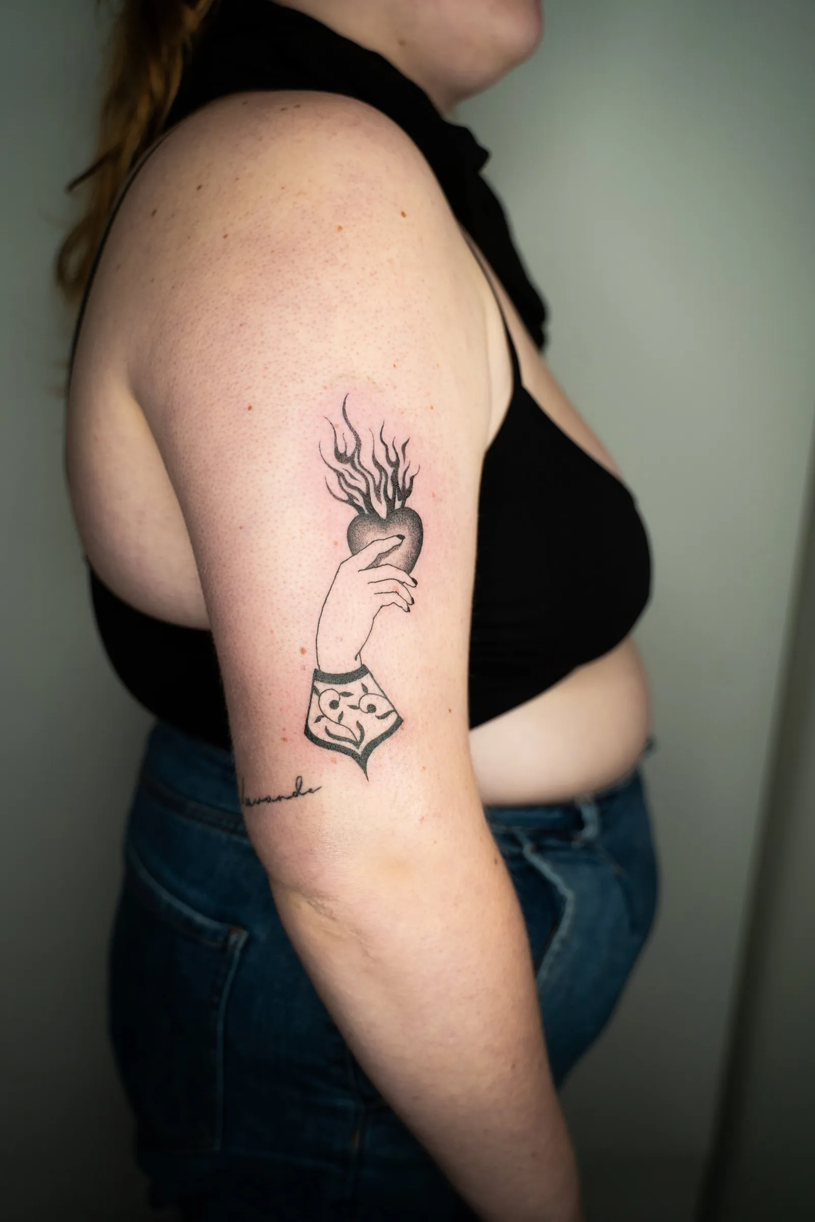 A hand in a lace cuff holding up a burning heart, tattooed on the back of an upper arm