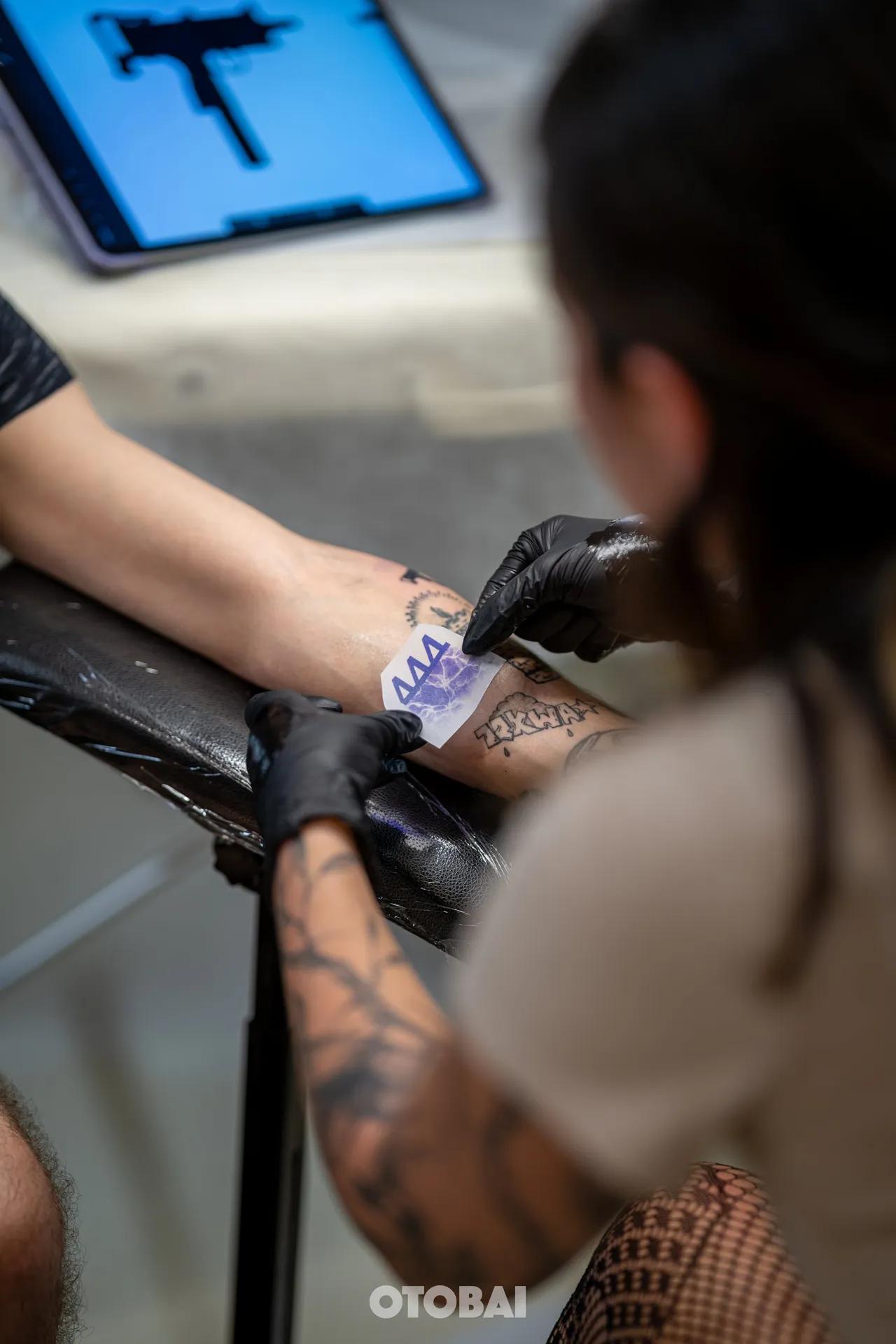Joy placing a stencil on a forearm, the design on a tablet beside her