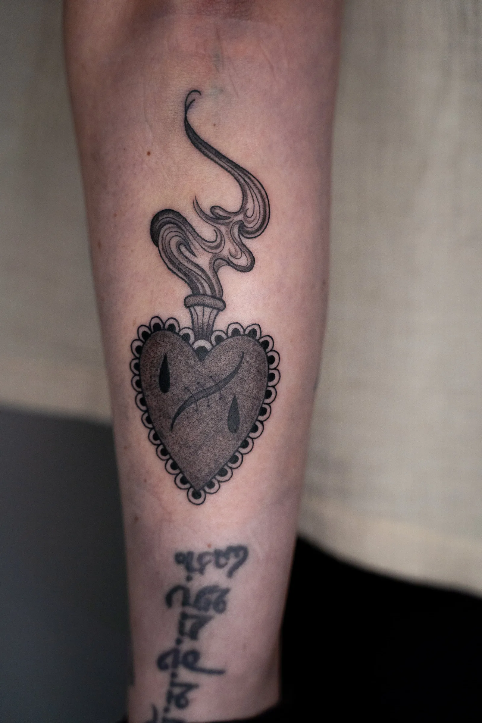 A sacred heart with a scalloped edge, teardrops and a plume of smoke, tattooed in black and grey on a shin