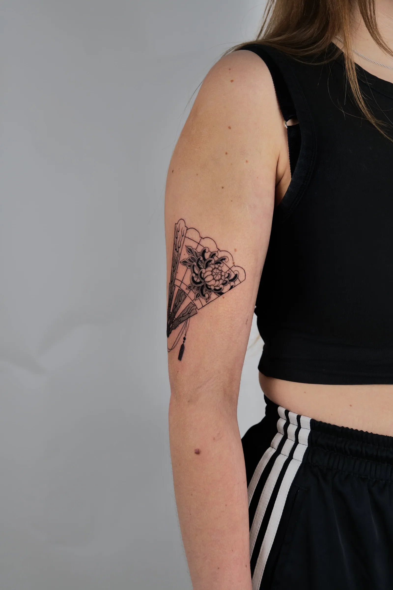 An open folding fan with a flower, tattooed in black and grey on an upper arm