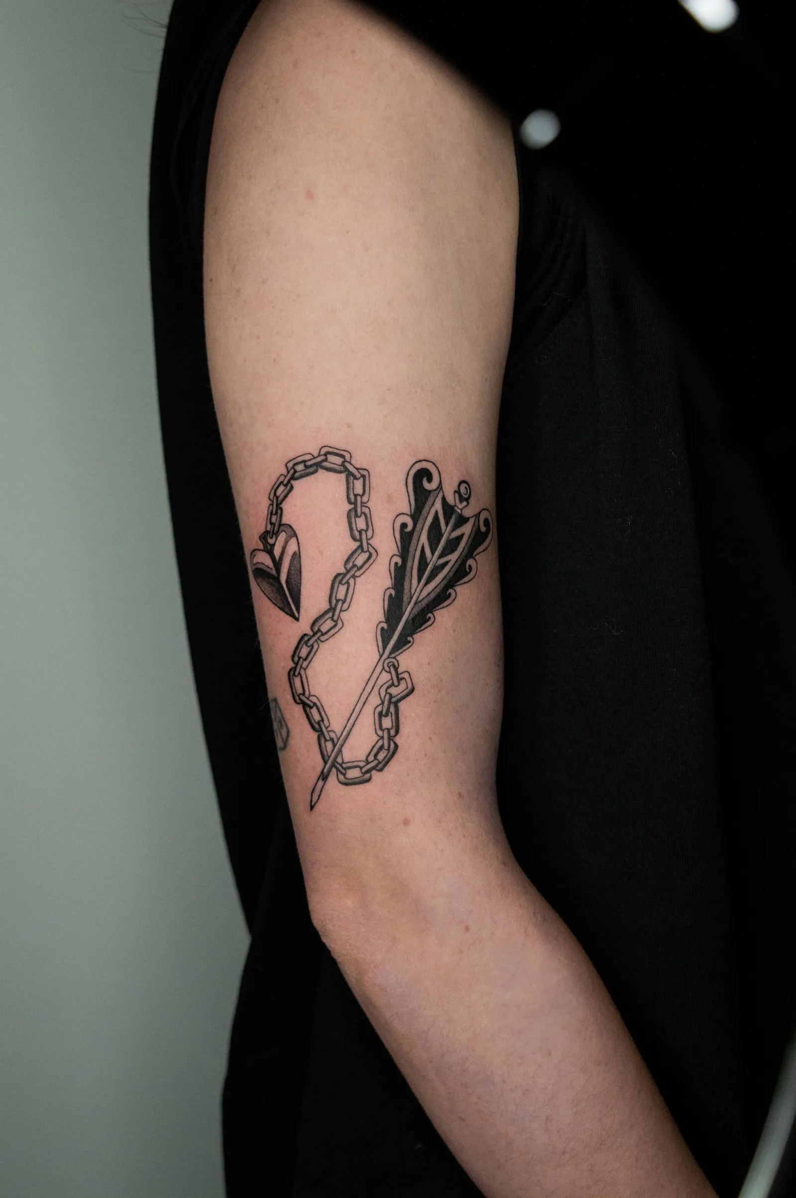 A looped chain with a feathered arrow and a small heart, tattooed on the back of an upper arm